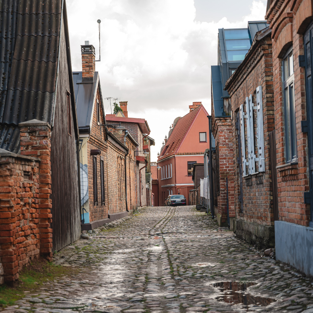 Siaura Kėdainių senamiesčio fotografija. Akmenimis grįsta gatvė, apsiniaukęs dangus, seni, rudų plytų namai iš abiejų gatvės pusių.